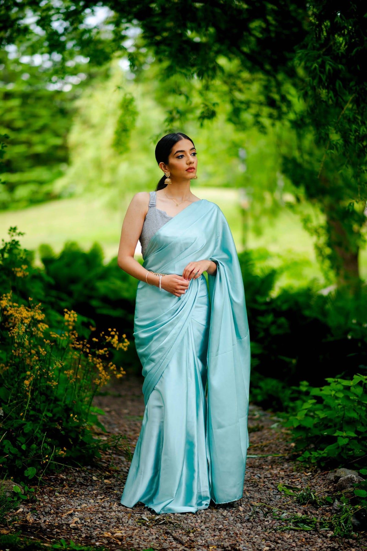 A model draped in an aqua blue satin silk saree, grey shimmer blouse and light blue saree shaper. The saree is pleated and  held with a waist chain.